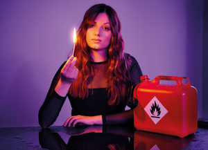 Lara Ermer sitzt mit einem brennenden Feuerzeug in der Hand neben einem offenen Benzinkanister in einem dunklen Raum mit lila Hintergrund