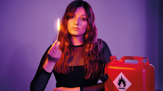 Lara Ermer sitzt mit einem brennenden Feuerzeug in der Hand neben einem offenen Benzinkanister in einem dunklen Raum mit lila Hintergrund